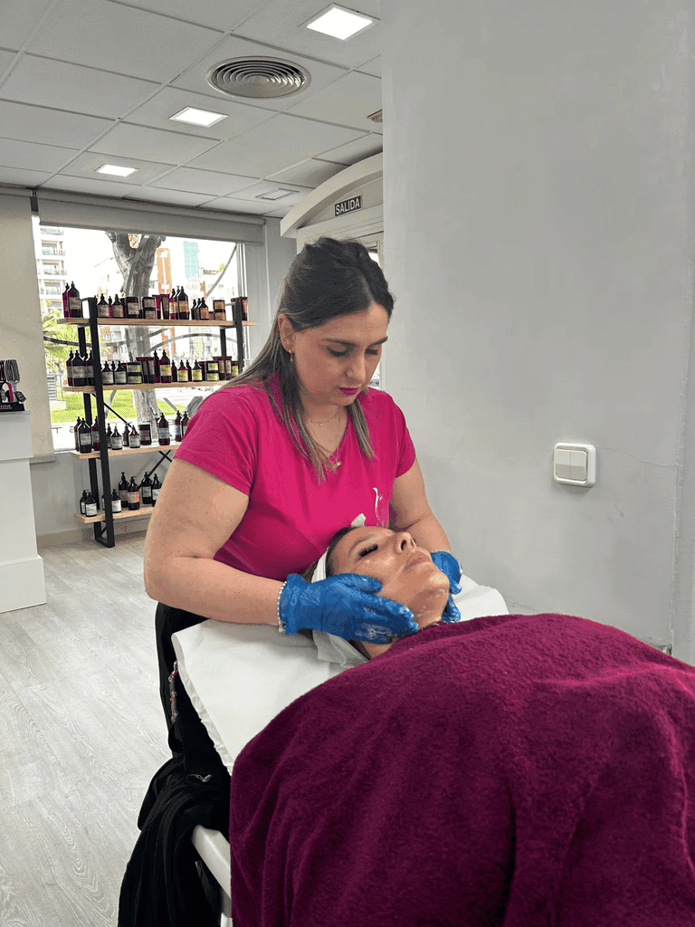 Trabajadora de Iderma con una camiseta rosa y guantes azules realiza un tratamiento facial a un cliente acostado en una camilla de tratamiento, cubierto con una manta marrón, en una sala de spa moderna y bien iluminada.  