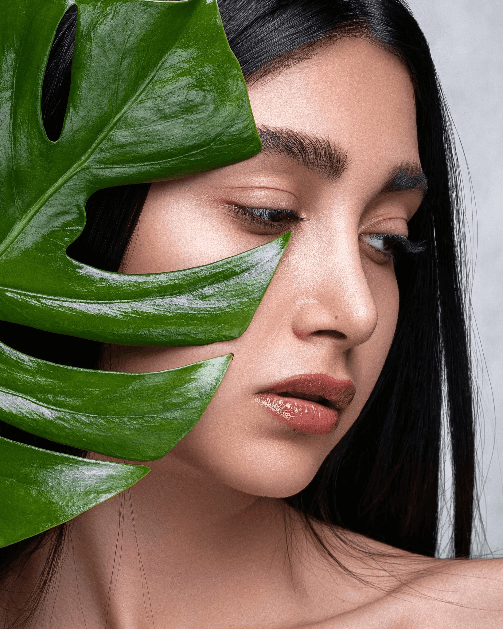 Una mujer de pelo largo y oscuro sostiene una gran hoja verde cerca de su rostro. Tiene labios brillantes y cejas definidas, y su mirada se dirige hacia abajo. El fondo es neutro, lo que la resalta a ella y a la hoja.