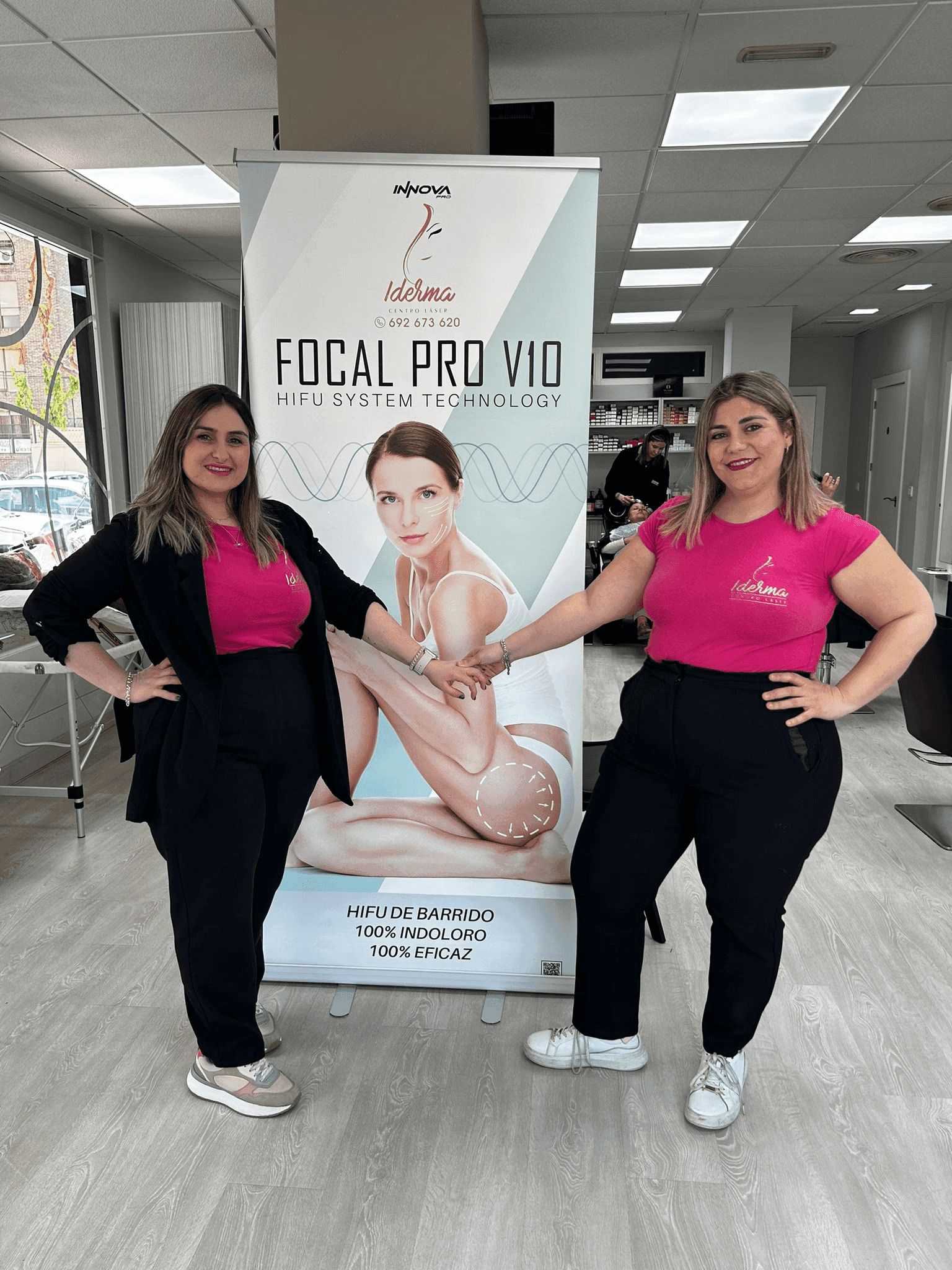 Dos mujeres sonrientes con uniformes de Iderma (camisetas rosas y pantalones negros) posan junto a un banner promocional de "FOCAL PRO V10 HIFU SYSTEM TECHNOLOGY". El banner muestra una imagen de una mujer desnuda y promociona un tratamiento HIFU.